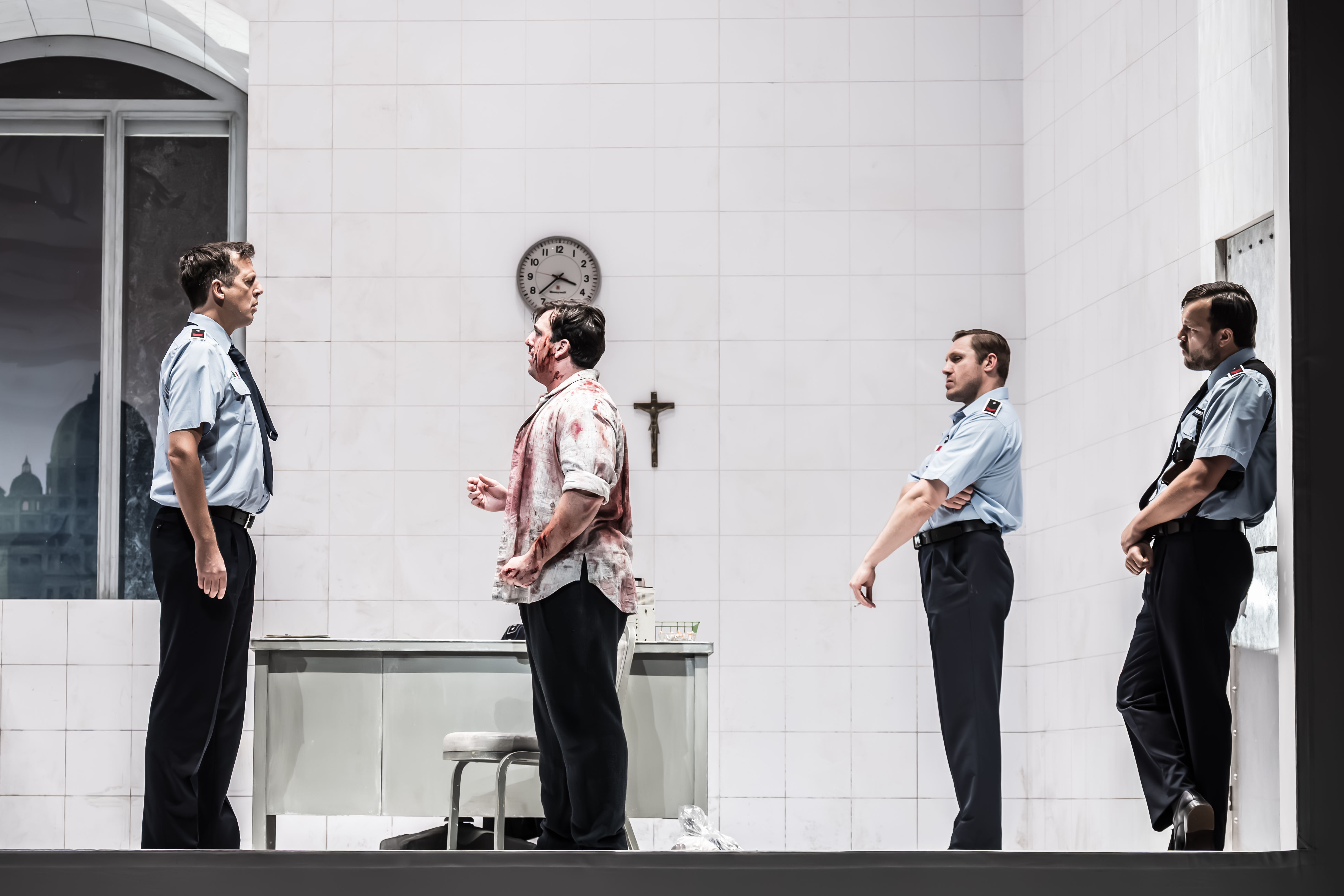 A performer wearing a blood stained shirt stands looking at a performer in a police uniform. Behind him are two other police officers. They are standing inside a brightly-lit stark white room with a crucafix and clock on the wall. (5 of 6)
