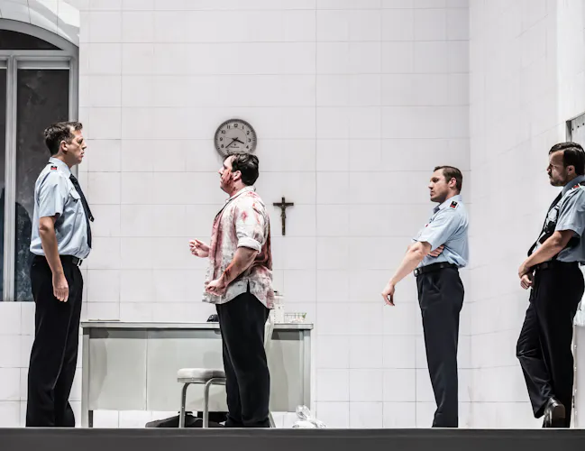A performer wearing a blood stained shirt stands looking at a performer in a police uniform. Behind him are two other police officers. They are standing inside a brightly-lit stark white room with a crucafix and clock on the wall. (5 of 6)