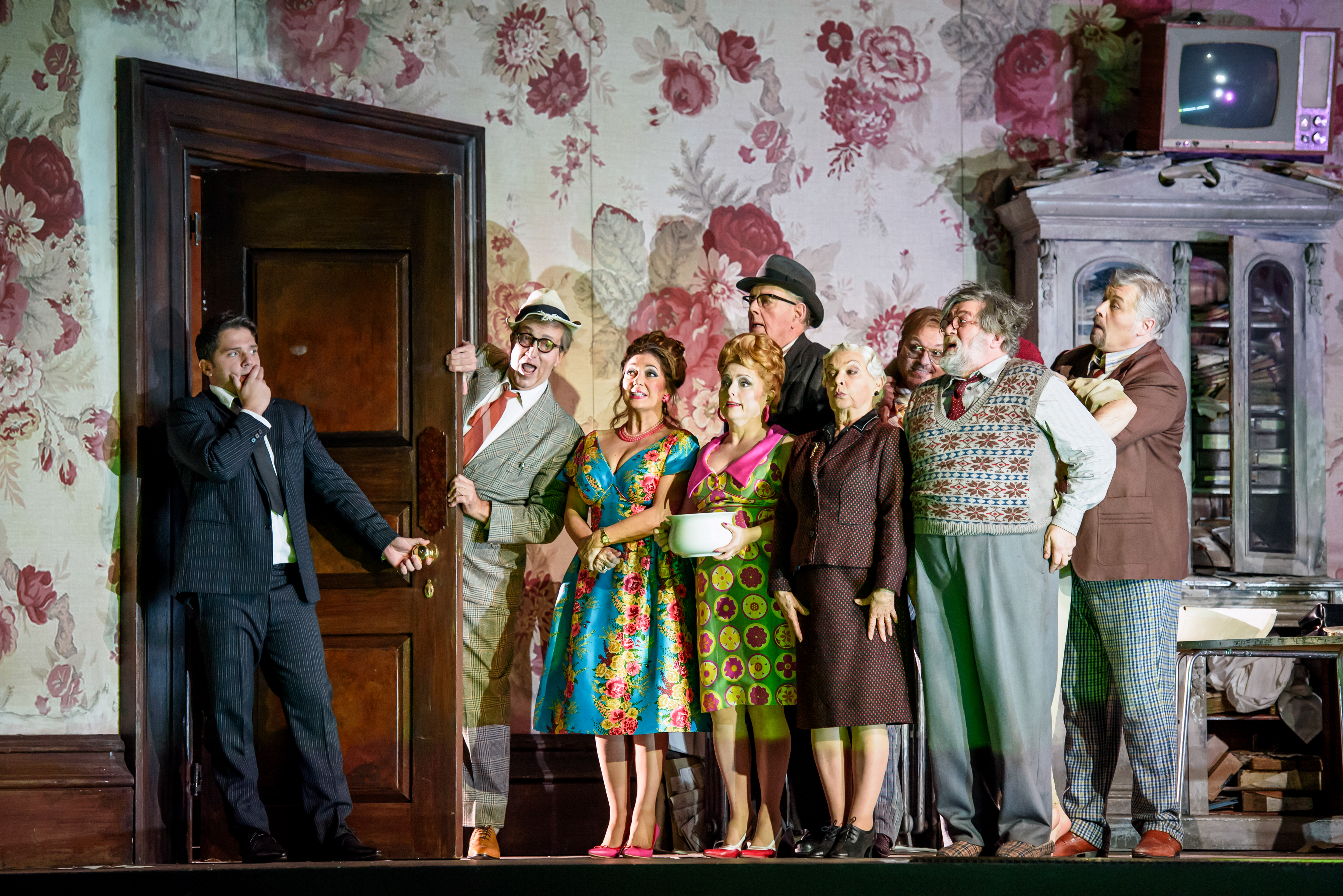 A group of performers stand alongside a door frame in a room with flowered wallpaper on stage. A performer wearing a suit has come through the door and holds onto the handle. 