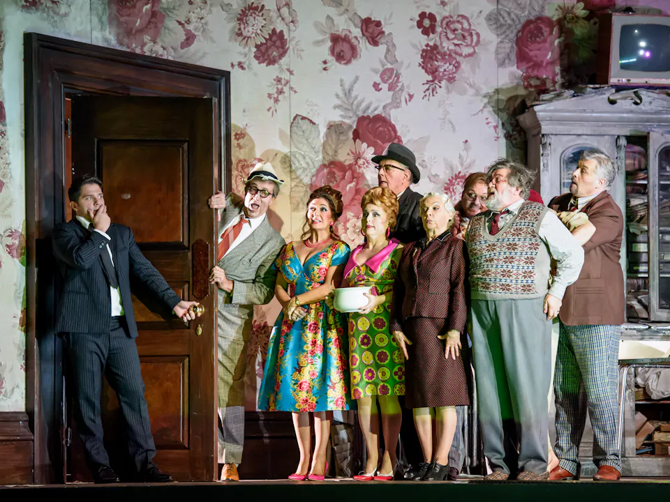 A group of performers stand alongside a door frame in a room with flowered wallpaper on stage. A performer wearing a suit has come through the door and holds onto the handle.