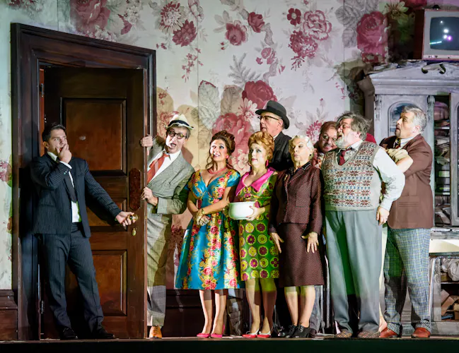 A group of performers stand alongside a door frame in a room with flowered wallpaper on stage. A performer wearing a suit has come through the door and holds onto the handle. (9 of 12)