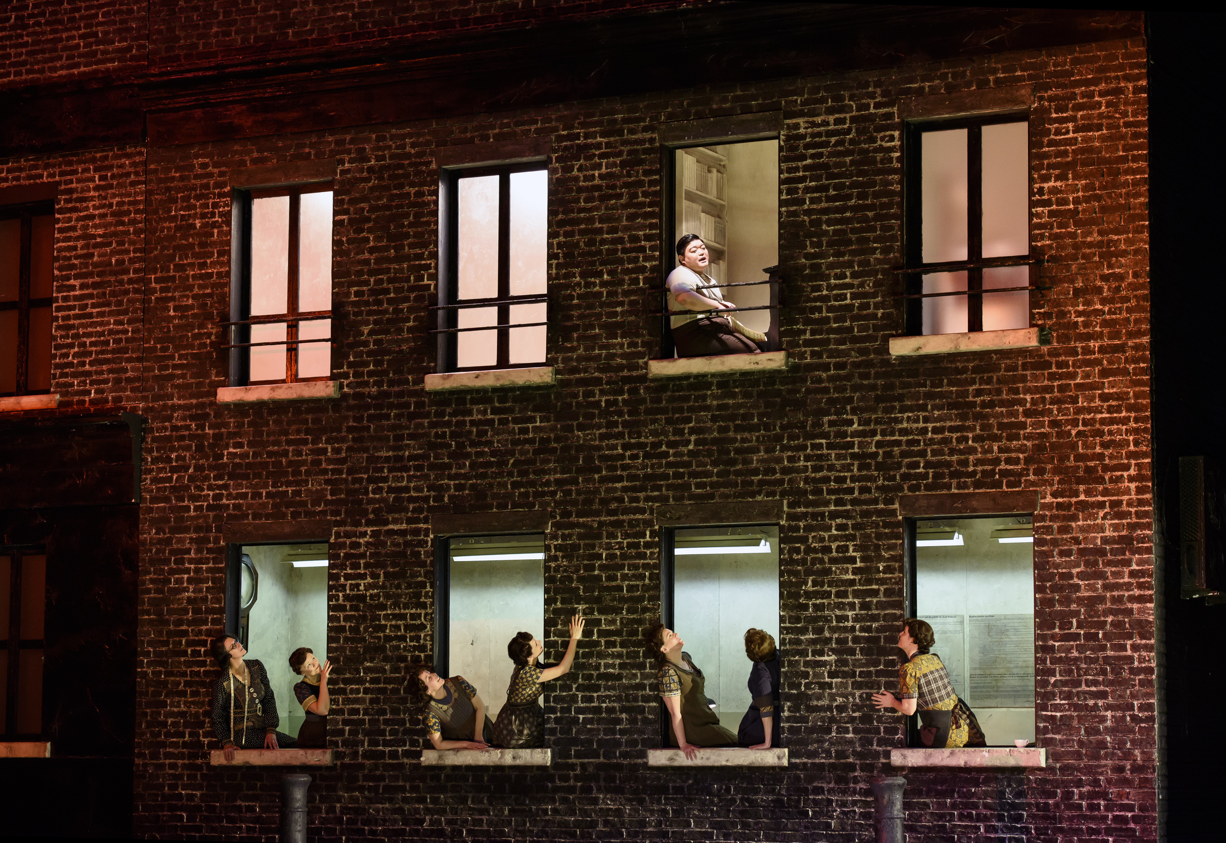 A red bricked house on stage with performers  leaning out of the bottom floor windows looking up towards one performer who sits at the window on the second floor. The stage is dark with lighting coming from inside the house. (1 of 6)