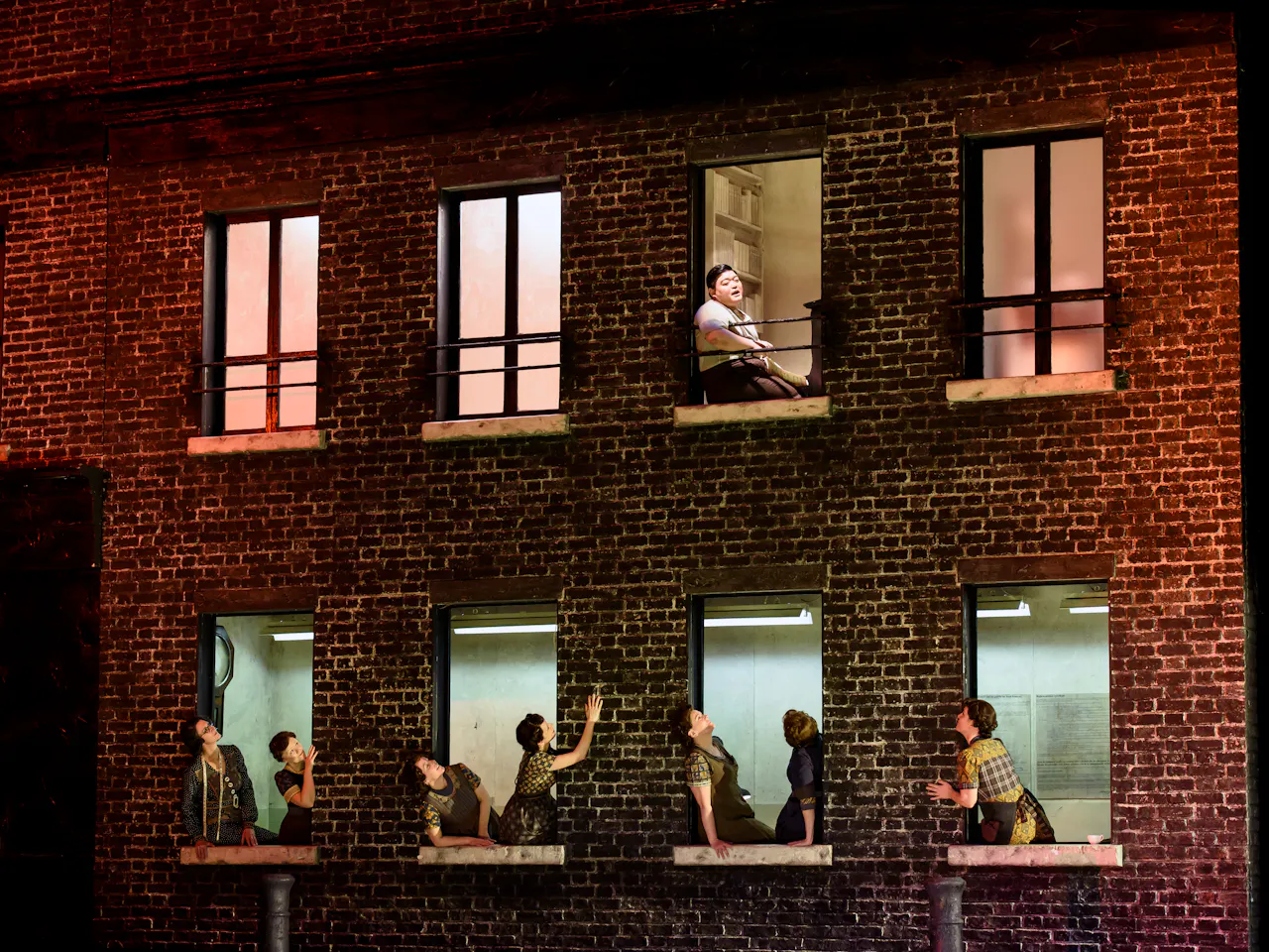 A red bricked house on stage with performers leaning out of the bottom floor windows looking up towards one performer who sits at the window on the second floor. The stage is dark with lighting coming from inside the house.