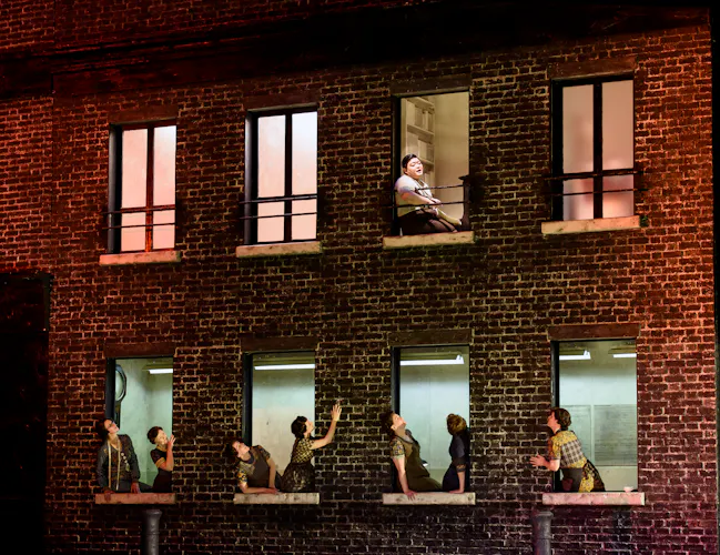 A red bricked house on stage with performers leaning out of the bottom floor windows looking up towards one performer who sits at the window on the second floor. The stage is dark with lighting coming from inside the house. (1 of 6)