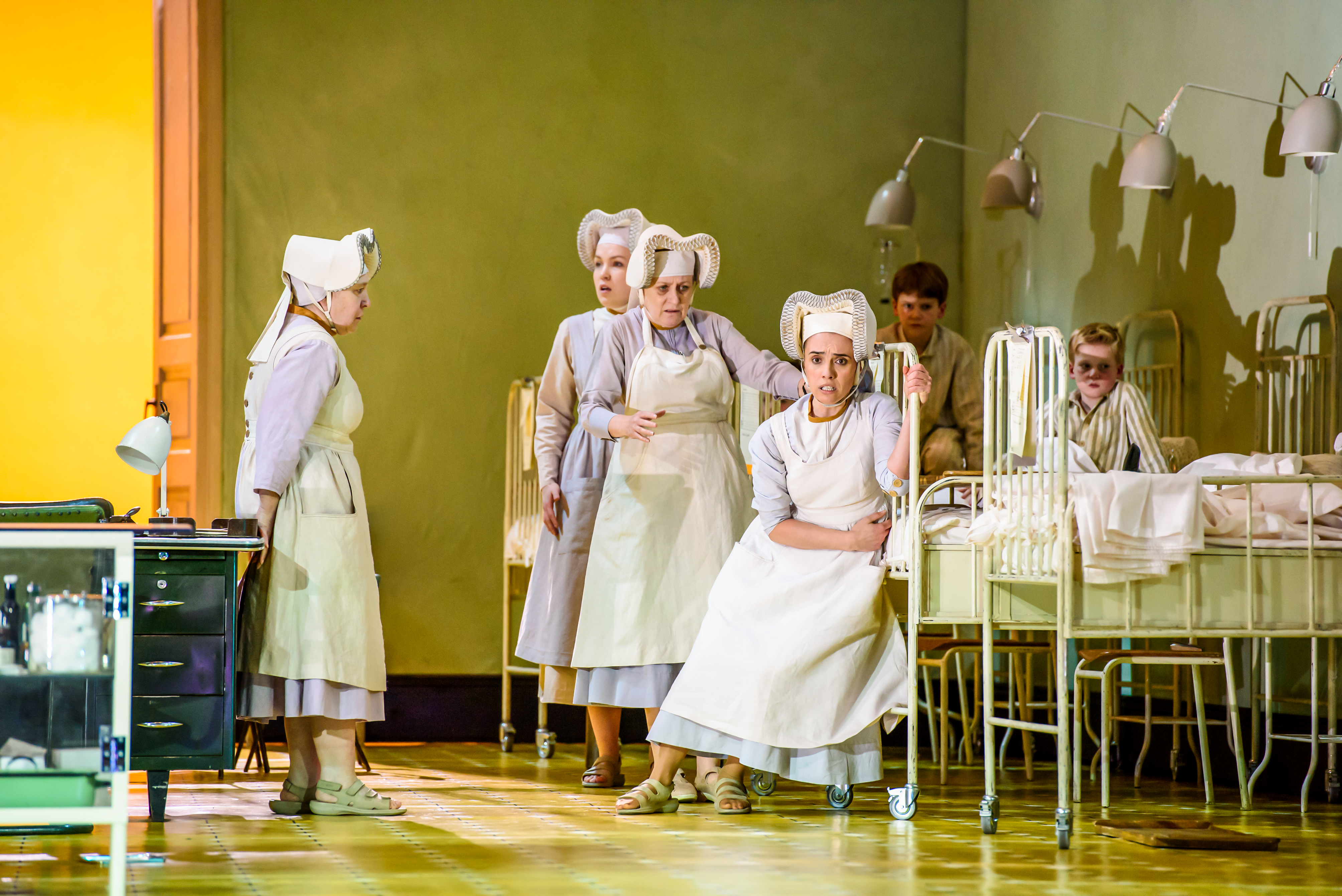 Several performers in nurses uniforms stand inside a hospital scene with metal framed beds.