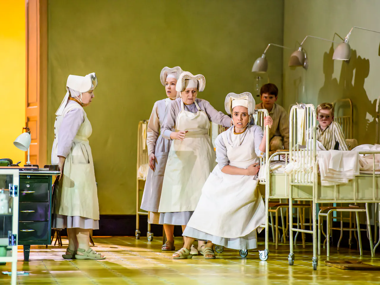 Several performers in nurses uniforms stand inside a hospital scene with metal framed beds.