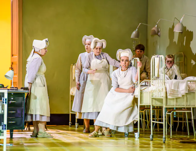 Several performers in nurses uniforms stand inside a hospital scene with metal framed beds. (2 of 6)