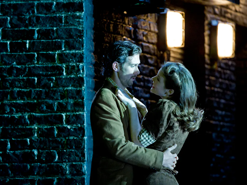 Two performers embrace passionately against a black painted brick wall with street lights.