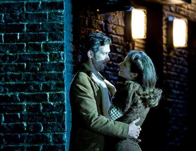 Two performers embrace passionately against a black painted brick wall with street lights. (4 of 6)