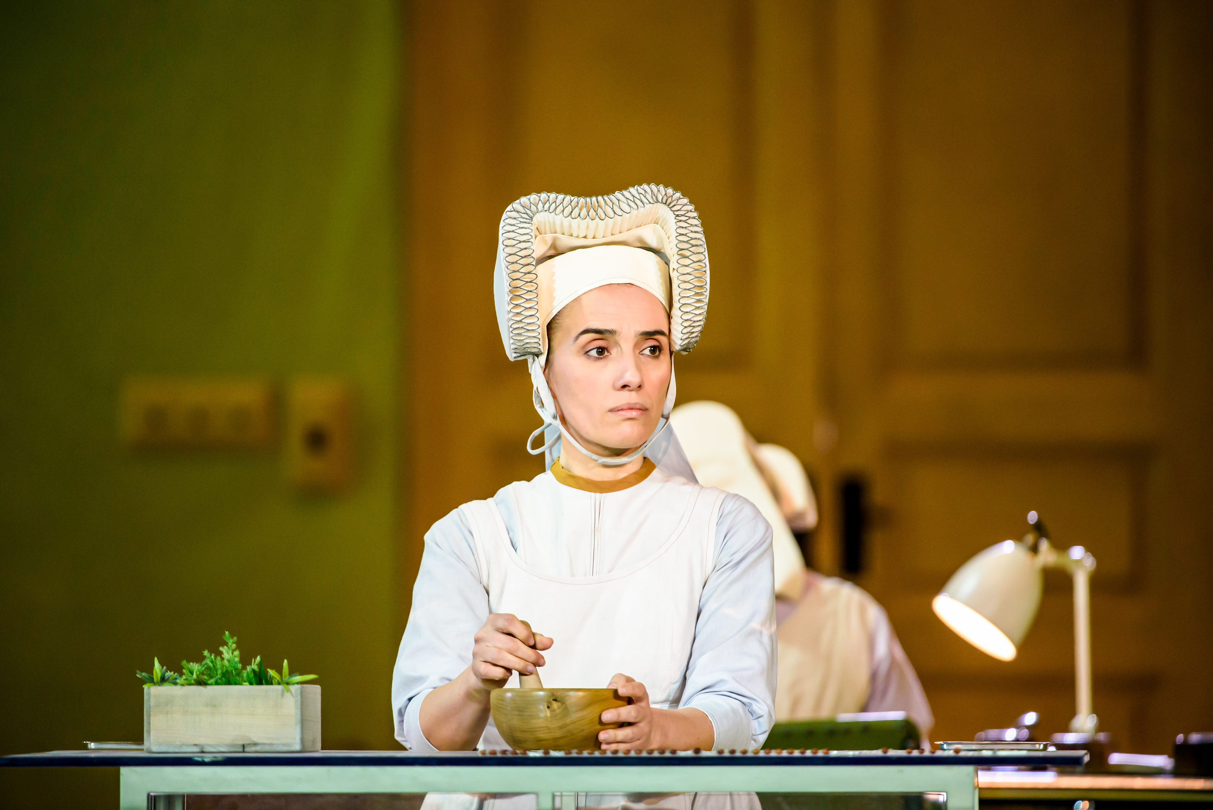 A performer wearing a medieval-style headdress sits at a table mixing a bowl. (3 of 6)