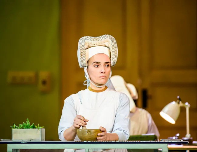 A performer wearing a medieval-style headdress sits at a table mixing a bowl. (3 of 6)