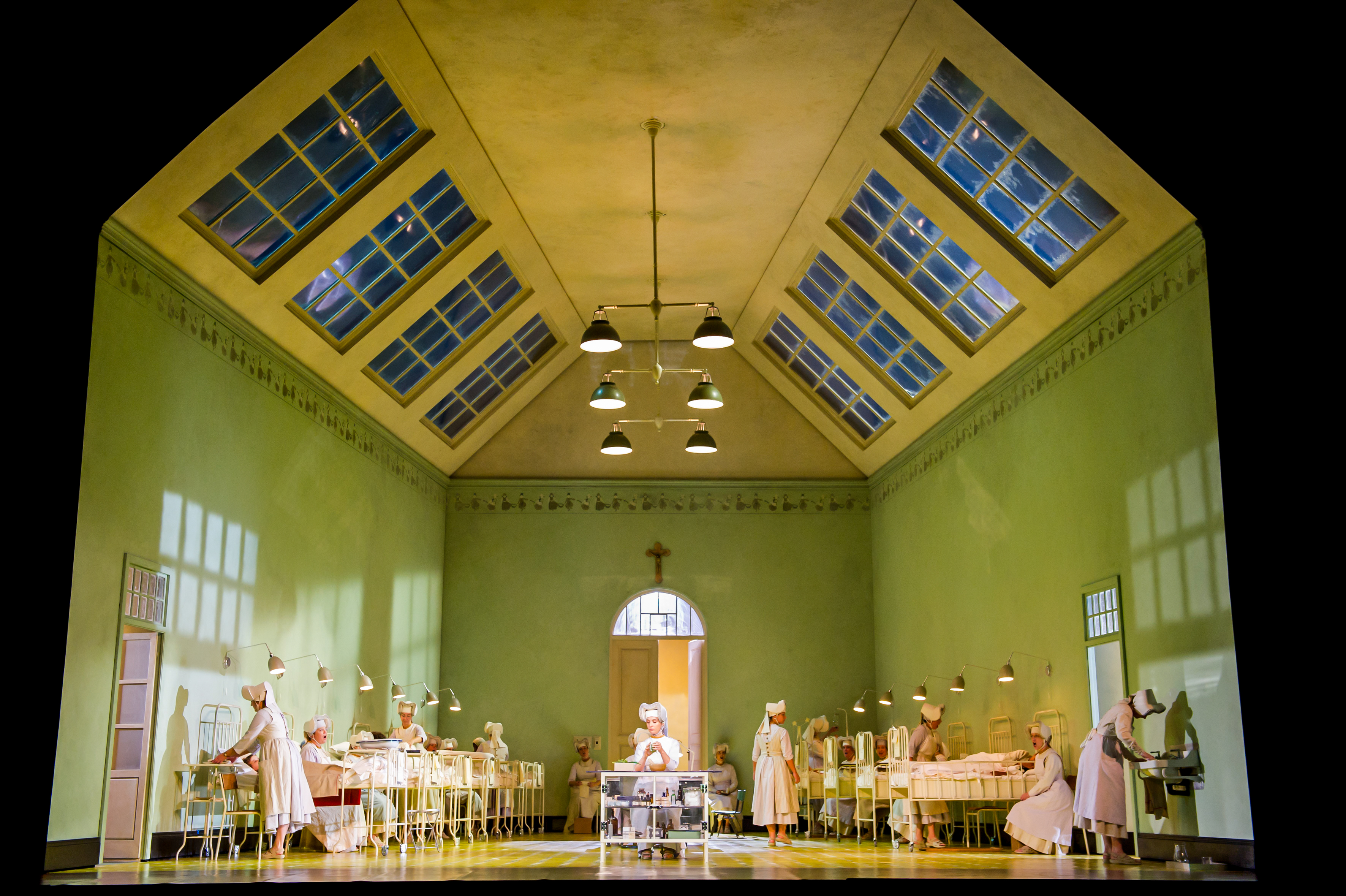 A hospital set on stage with green walls and a yellow ceiling with skylights. Several metal beds are either side of the room with performers in nurses uniforms moving throughout. (5 of 6)