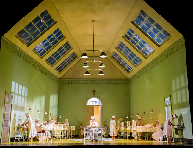 A hospital set on stage with green walls and a yellow ceiling with skylights. Several metal beds are either side of the room with performers in nurses uniforms moving throughout. (5 of 6)