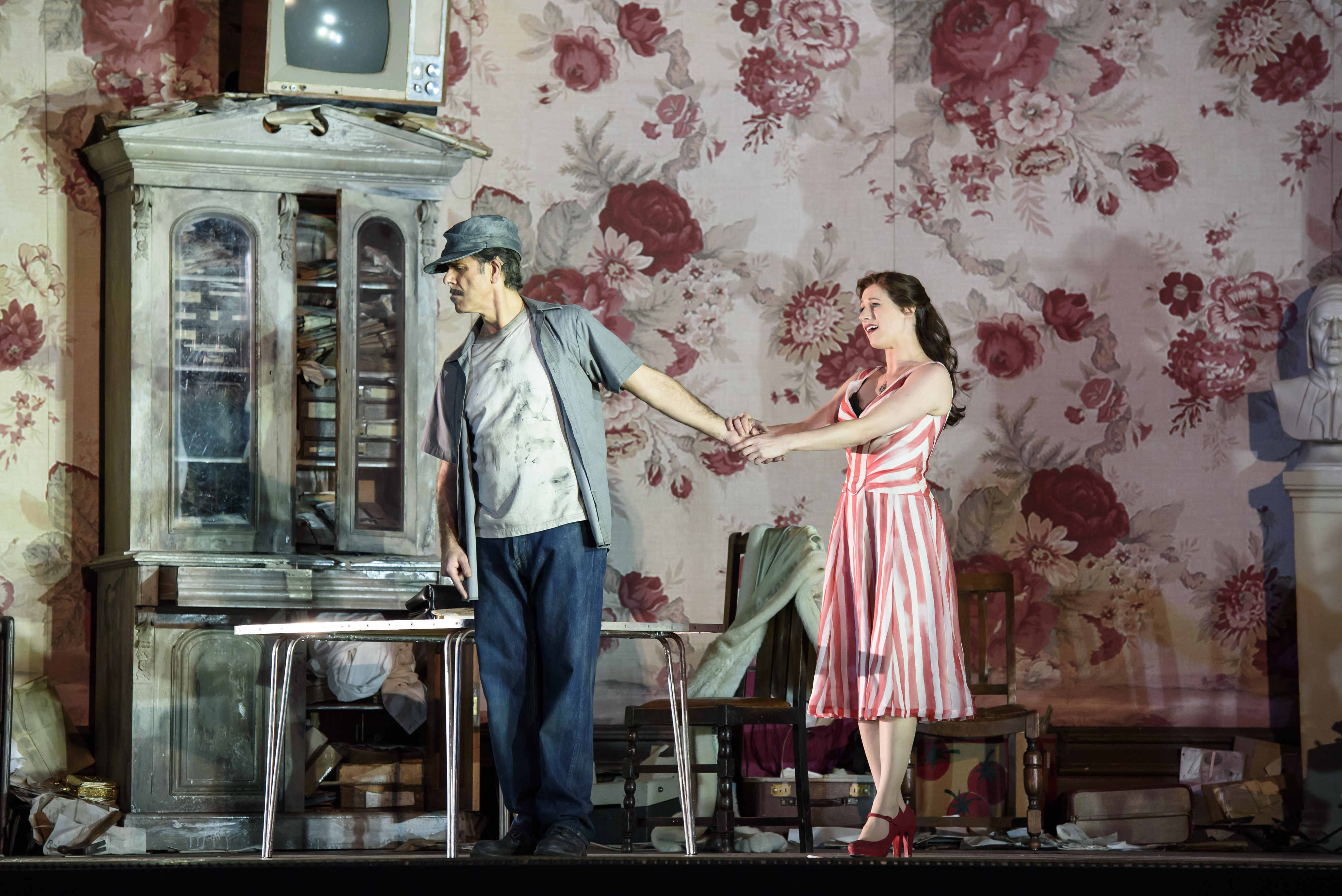 A performer wearing a pink adn white striped dress holds the hand of another performer who has their back to her. They stand inside a room on stage with red and pink wallpaper and living room furniture. (6 of 6)