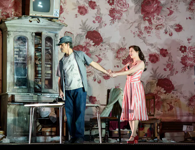 A performer wearing a pink adn white striped dress holds the hand of another performer who has their back to her. They stand inside a room on stage with red and pink wallpaper and living room furniture. (6 of 6)