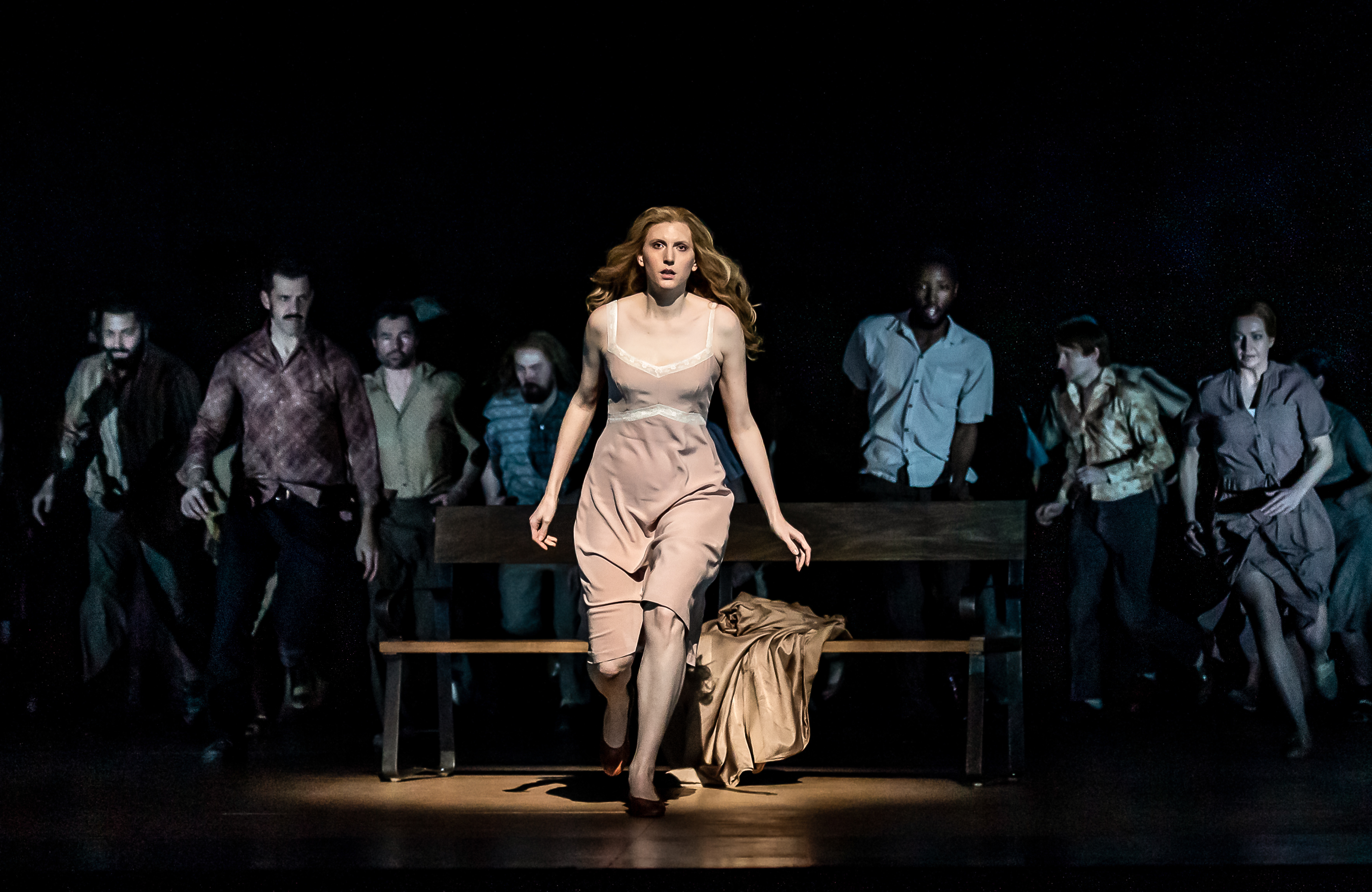 A performer wearing a beige dress is lit from above on stage and stands in front of a park bench with a beige coat strewn across. They are in movement as though running towards the audience. Behind them, only slightly visible, are a group of performers.