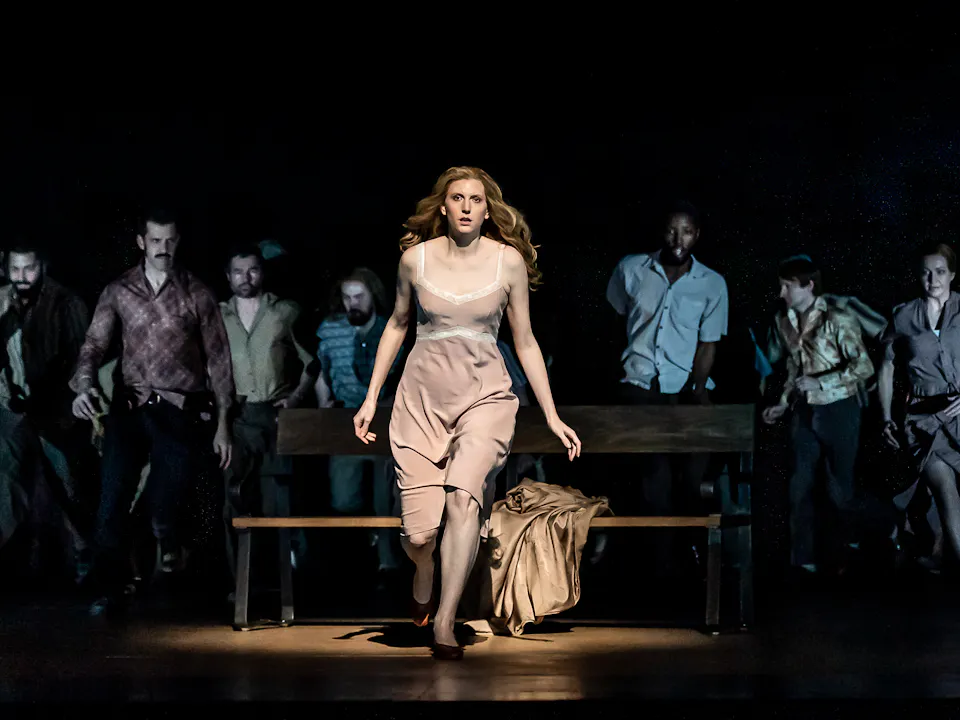 A performer wearing a beige dress is lit from above on stage and stands in front of a park bench with a beige coat strewn across. They are in movement as though running towards the audience. Behind them, only slightly visible, are a group of performers.
