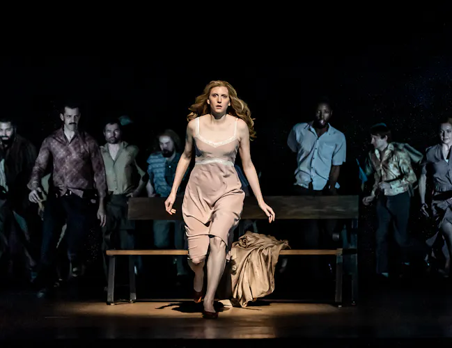 A performer wearing a beige dress is lit from above on stage and stands in front of a park bench with a beige coat strewn across. They are in movement as though running towards the audience. Behind them, only slightly visible, are a group of performers. (10 of 12)