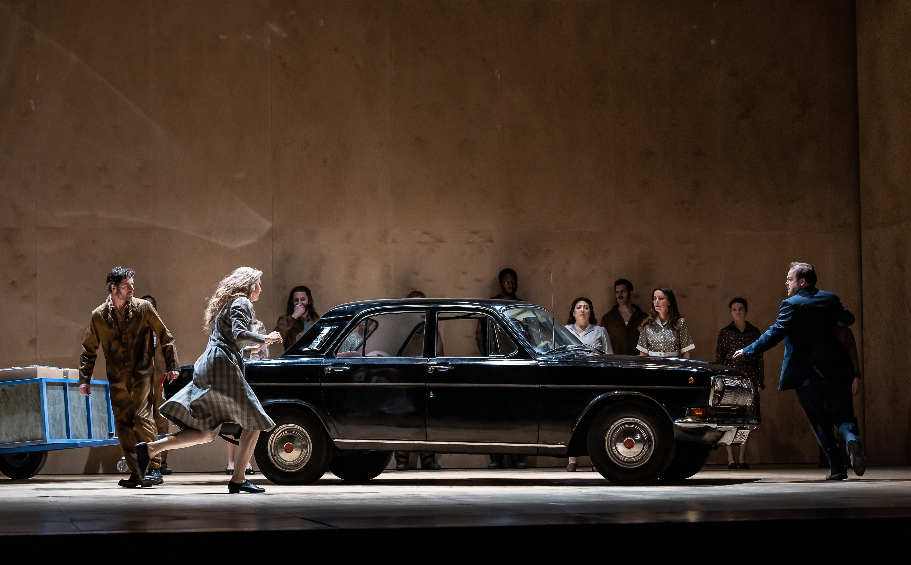 A vintage black car with a blue metal toe truck sits on stage with several performers moving around it. (1 of 7)