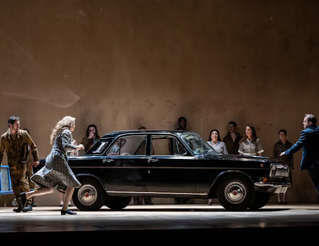 A vintage black car with a blue metal toe truck sits on stage with several performers moving around it. (1 of 7)
