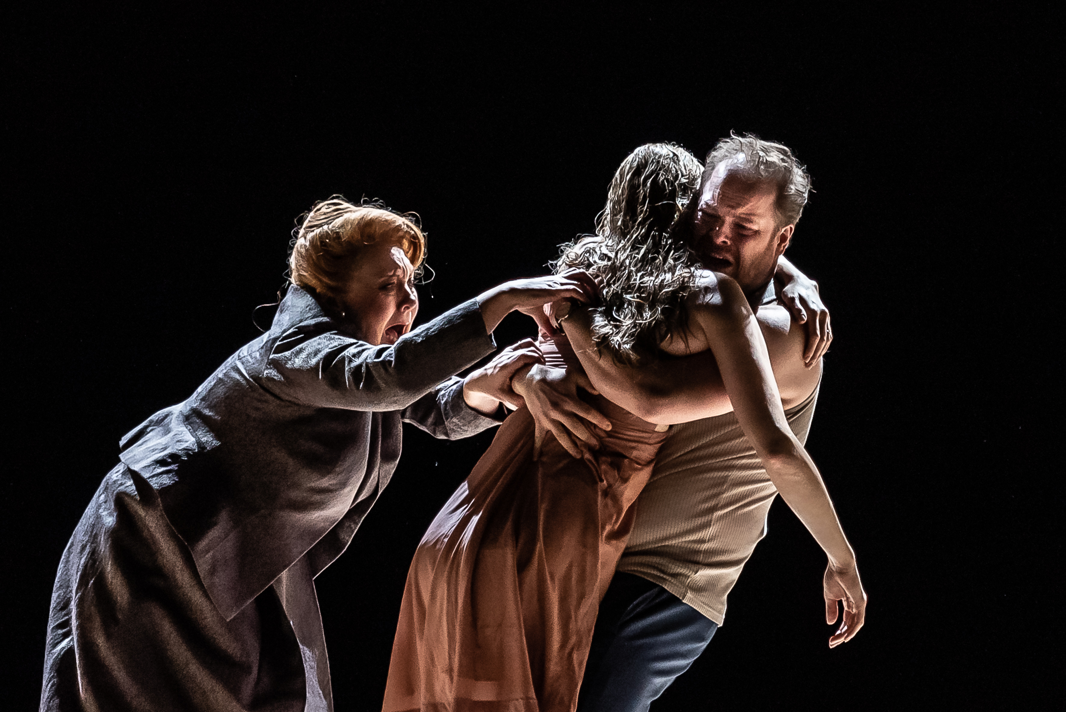 Two performers embrace, with one appearing limp with their arms draped down. Another performer desperately grabs to the limp performer. (2 of 7)