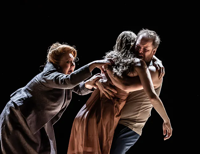 Two performers embrace, with one appearing limp with their arms draped down. Another performer desperately grabs to the limp performer. (2 of 7)