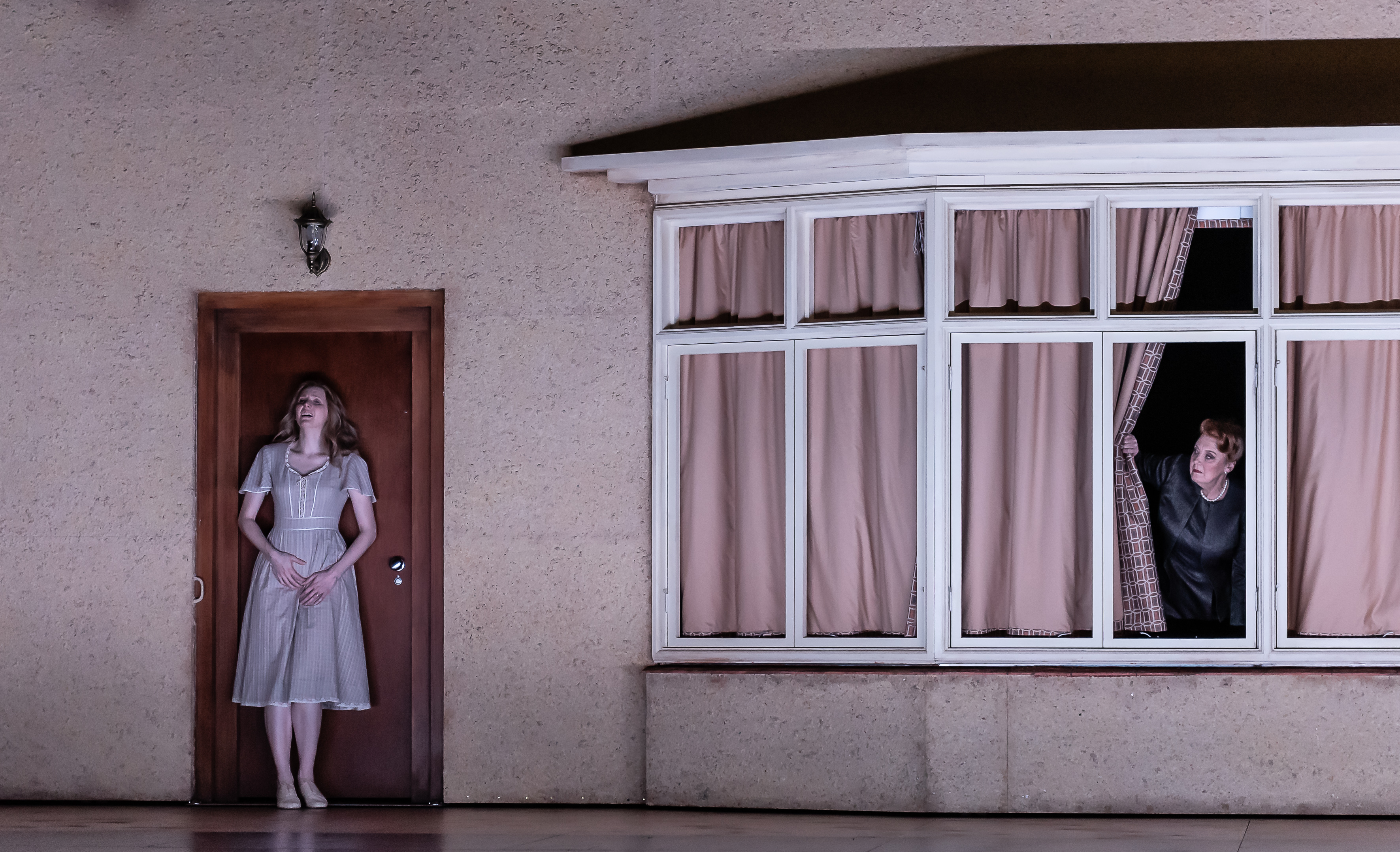 A performer stands outside the front door of a house on stage. Another performer peeps from the curtains from inside the house. (3 of 7)