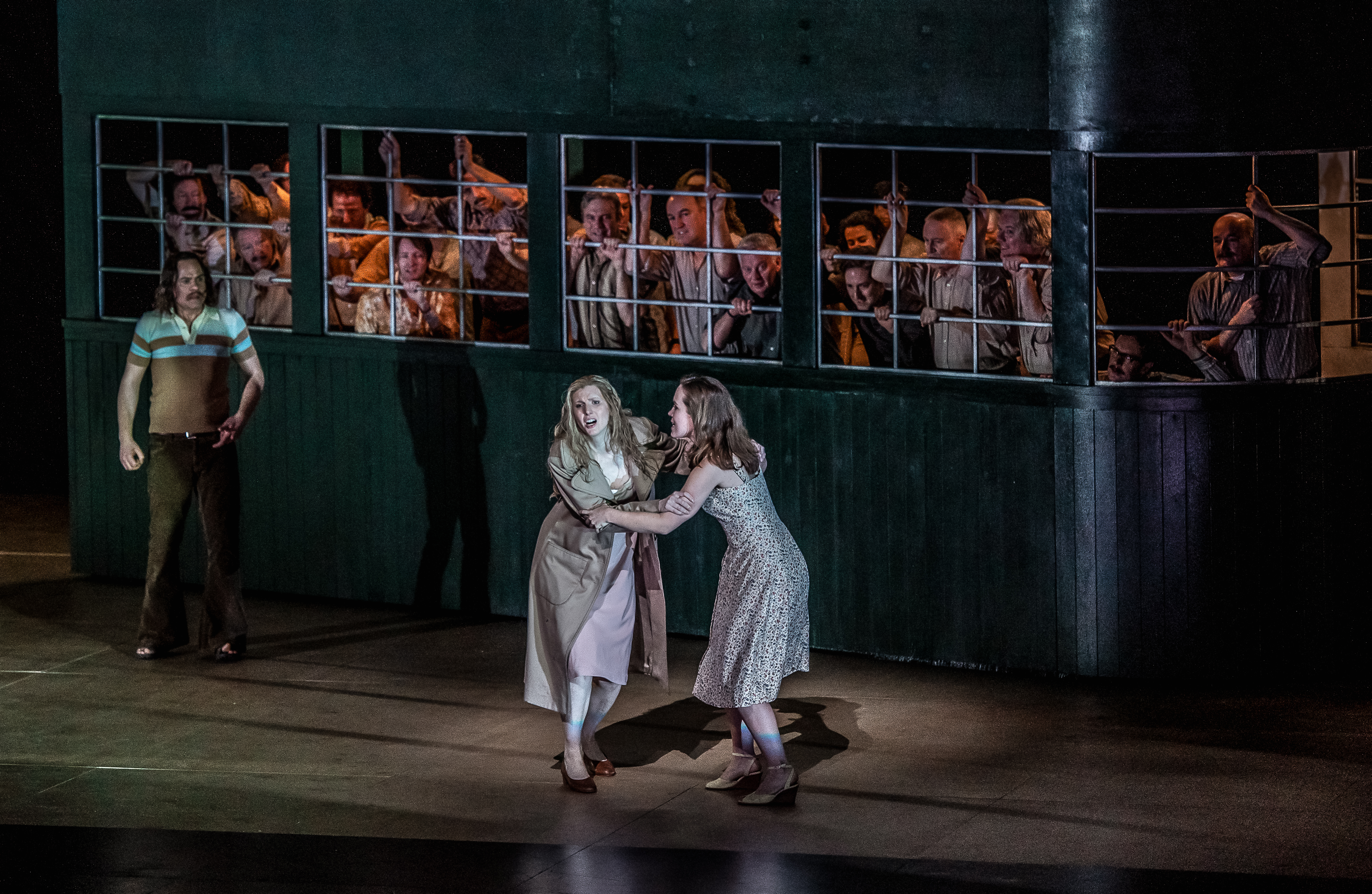Two performers hang onto each other as they sing. Behind them is a house with a large window that is filled with many performers looking out. (4 of 7)