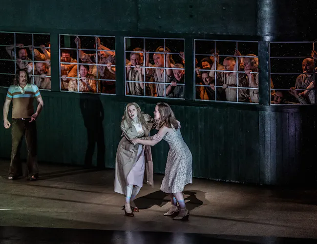 Two performers hang onto each other as they sing. Behind them is a house with a large window that is filled with many performers looking out. (4 of 7)