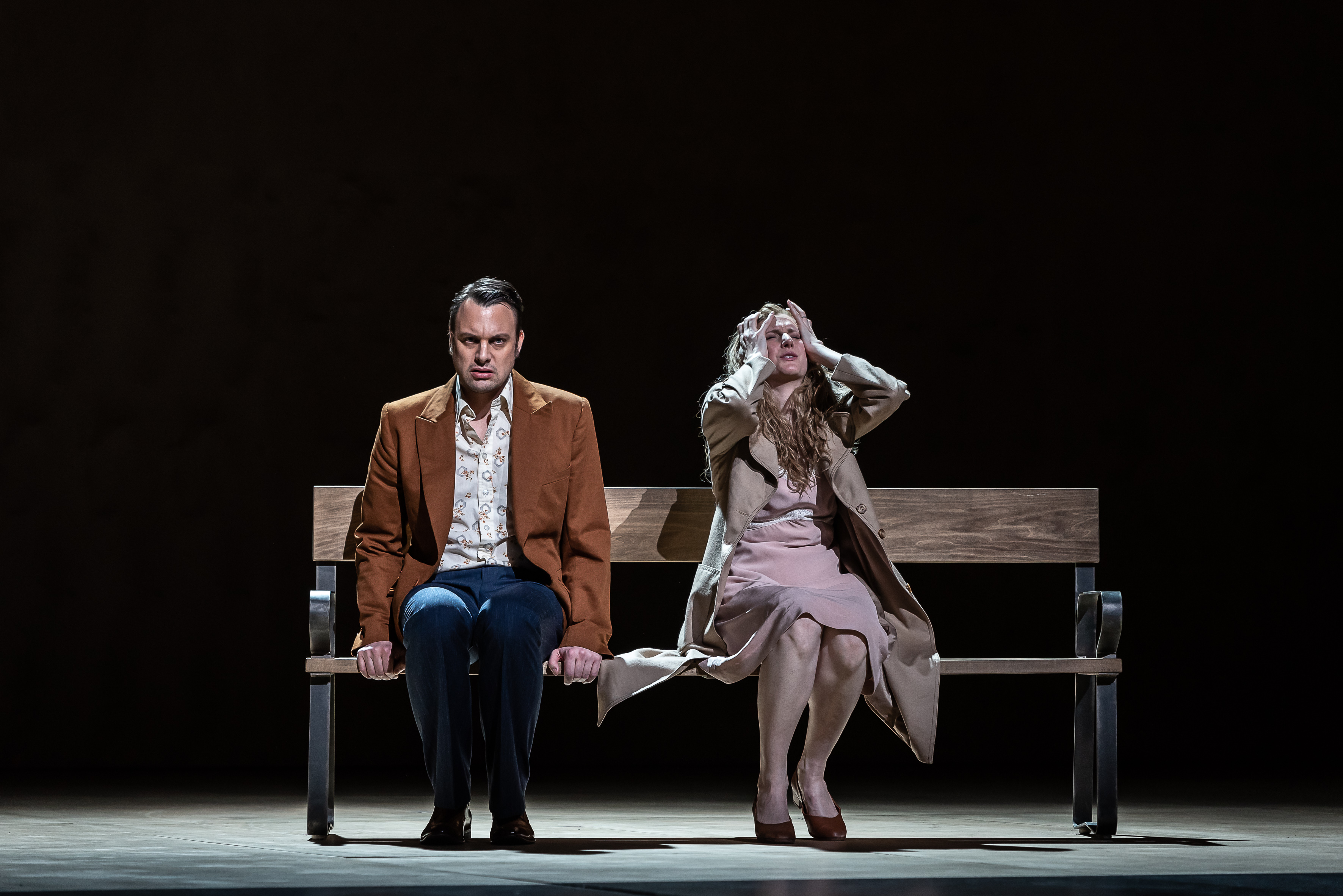 Two performers sit on a park bench on a dark stage. One holds their arms up to their head in distress. (5 of 7)