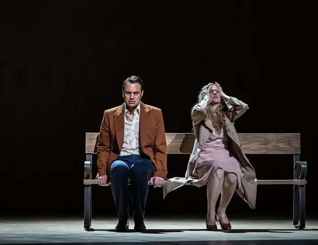 Two performers sit on a park bench on a dark stage. One holds their arms up to their head in distress. (5 of 7)