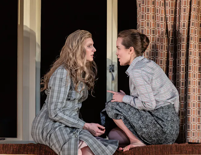 Two performers sit on a sofa facing one another by a window with retro curtains. (6 of 7)