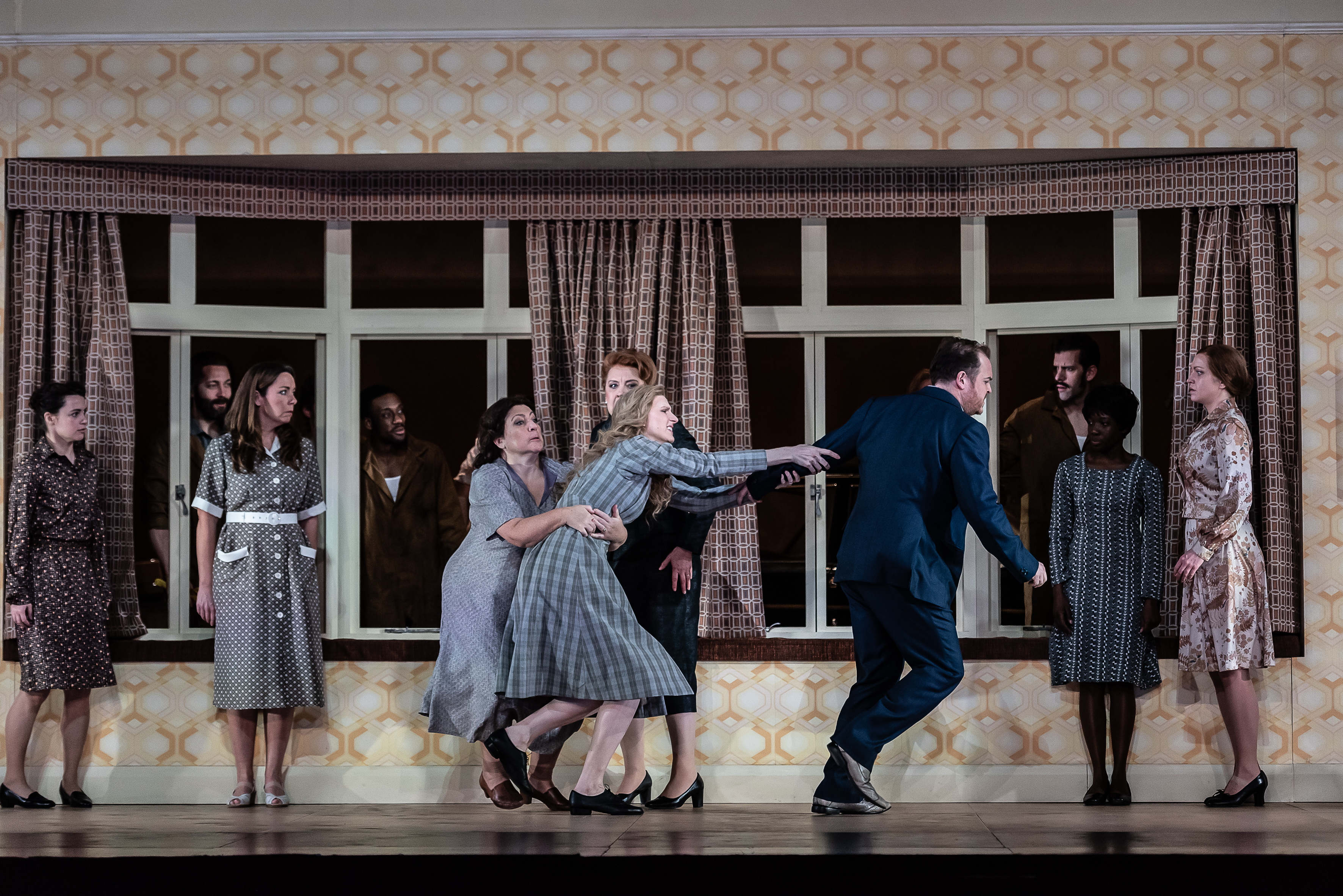A performer wearing a check dress lunges towards a person in a suit and is held back by another woman in a grey dress. There are several people around watching on. They are in front of a window with retro curtains and yellow retro wallpaper on stage. (7 of 7)