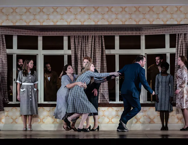 A performer wearing a check dress lunges towards a person in a suit and is held back by another woman in a grey dress. There are several people around watching on. They are in front of a window with retro curtains and yellow retro wallpaper on stage. (7 of 7)