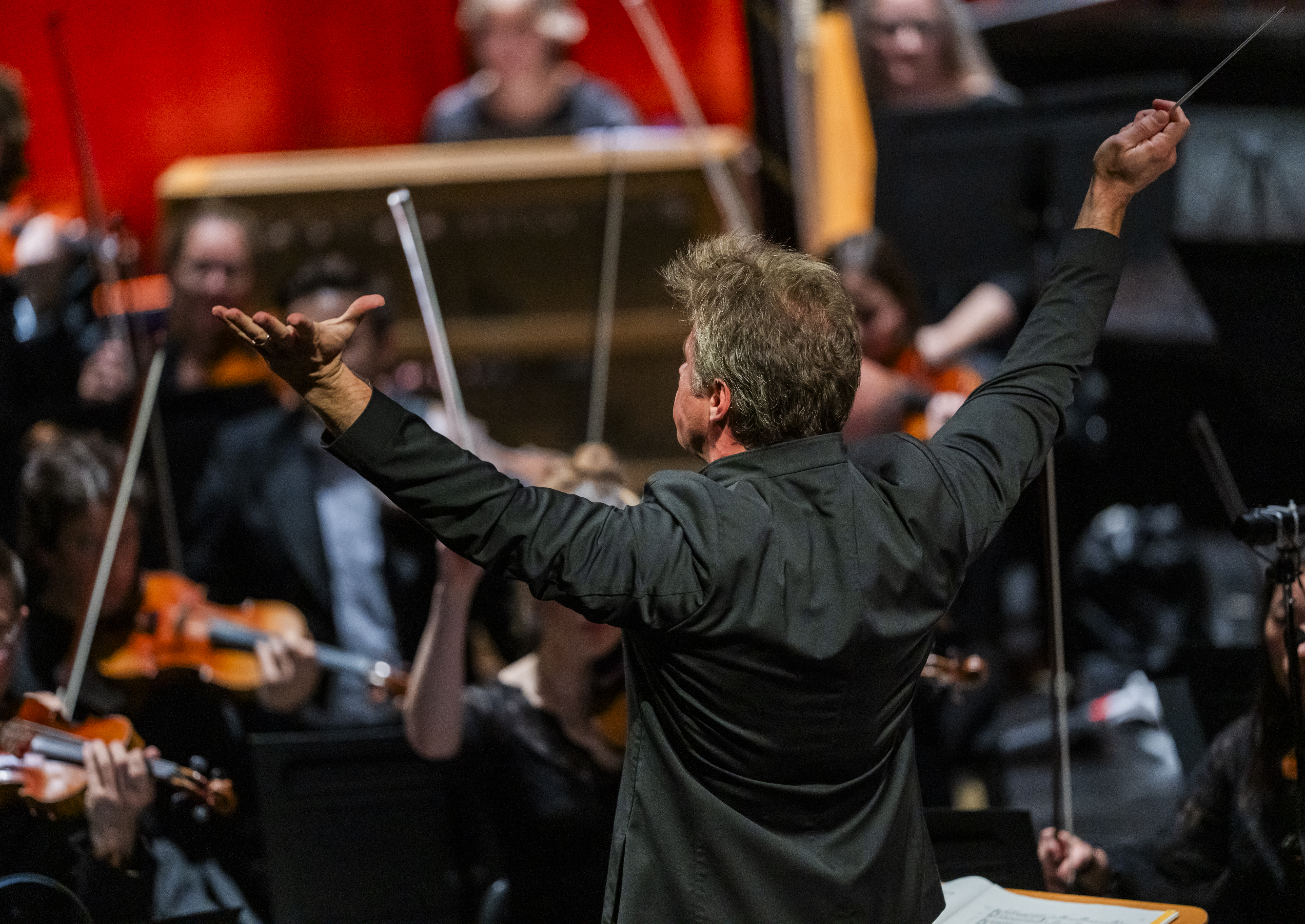 Music Director of The Royal Opera, Jakub Hrůša, conducts several members of an orchestra in a brightly lit room. He holds his arms upwards with a baton in his right hand. (1 of 4)