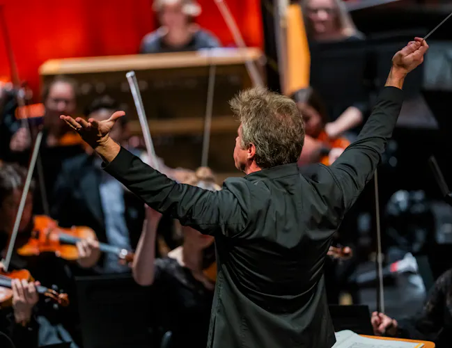 Music Director of The Royal Opera, Jakub Hrůša, conducts several members of an orchestra in a brightly lit room. He holds his arms upwards with a baton in his right hand. (1 of 4)
