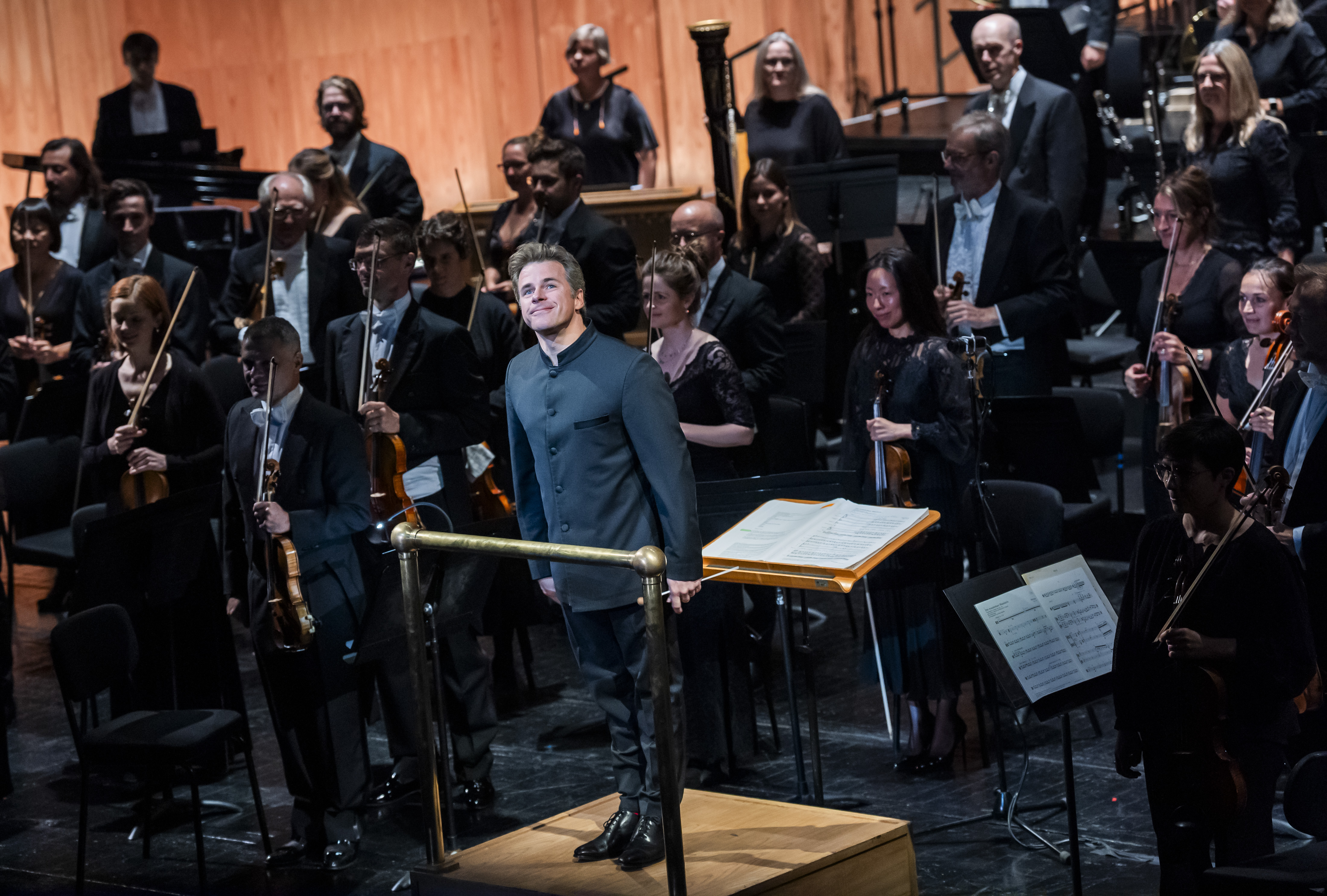 Music Director of The Royal Opera, Jakub Hrůša, stands in front of an orchestra in a conductor pit facing the audience.
