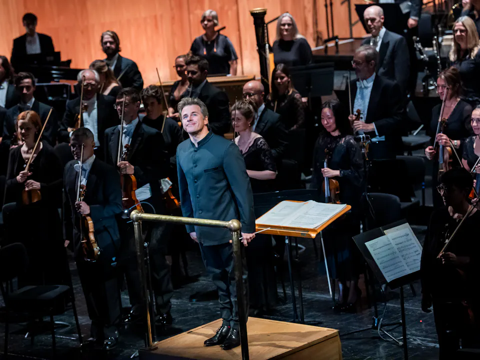 Music Director of The Royal Opera, Jakub Hrůša, stands in front of an orchestra in a conductor pit facing the audience.