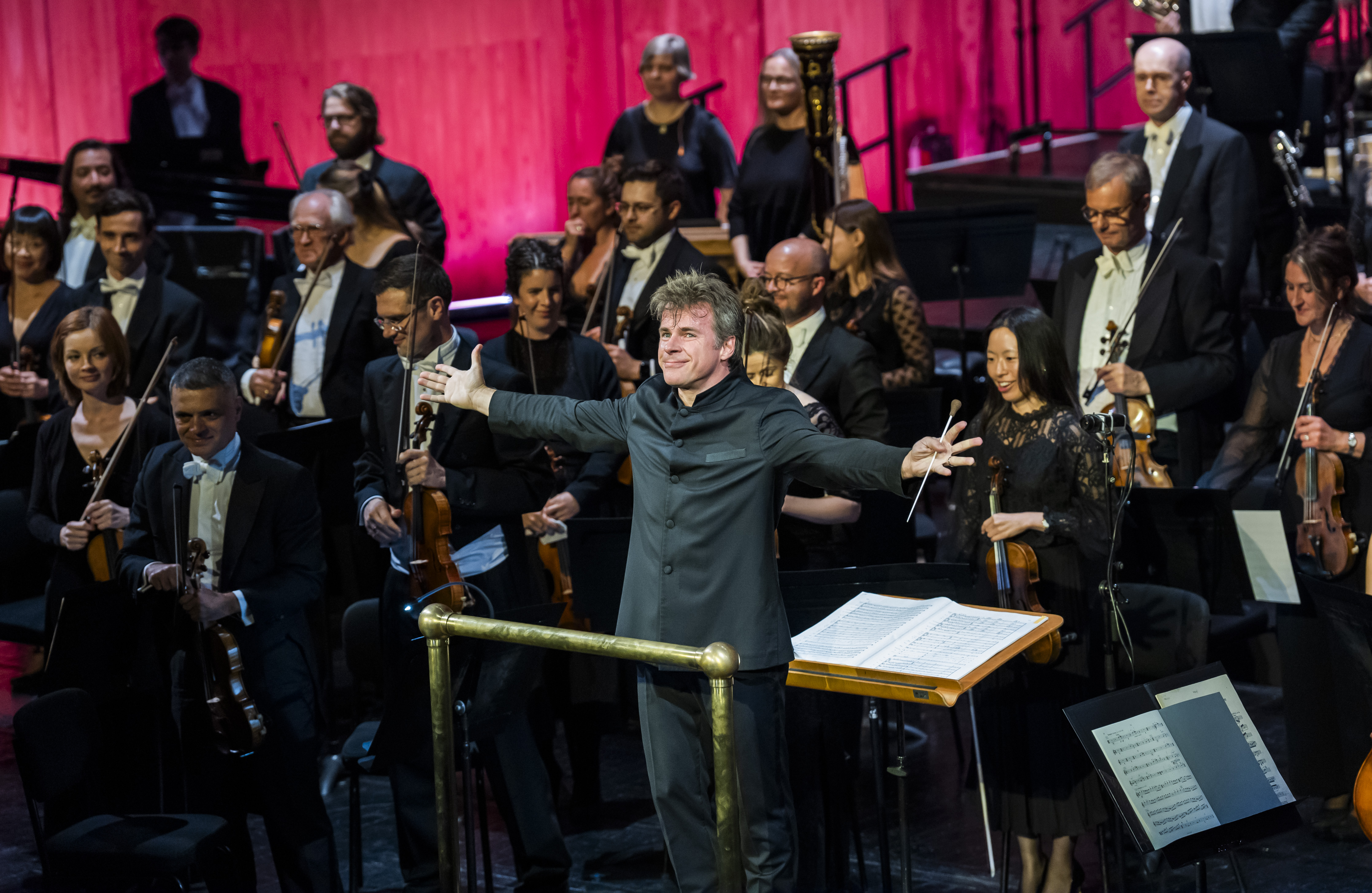 Music Director of The Royal Opera, Jakub Hrůša, stands in front of an orchestra in a conductor pit facing the audience with both his arms stretched outwards. He holds a baton in his left hand. (3 of 4)