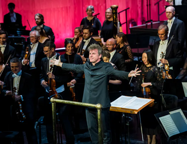 Music Director of The Royal Opera, Jakub Hrůša, stands in front of an orchestra in a conductor pit facing the audience with both his arms stretched outwards. He holds a baton in his left hand. (3 of 4)