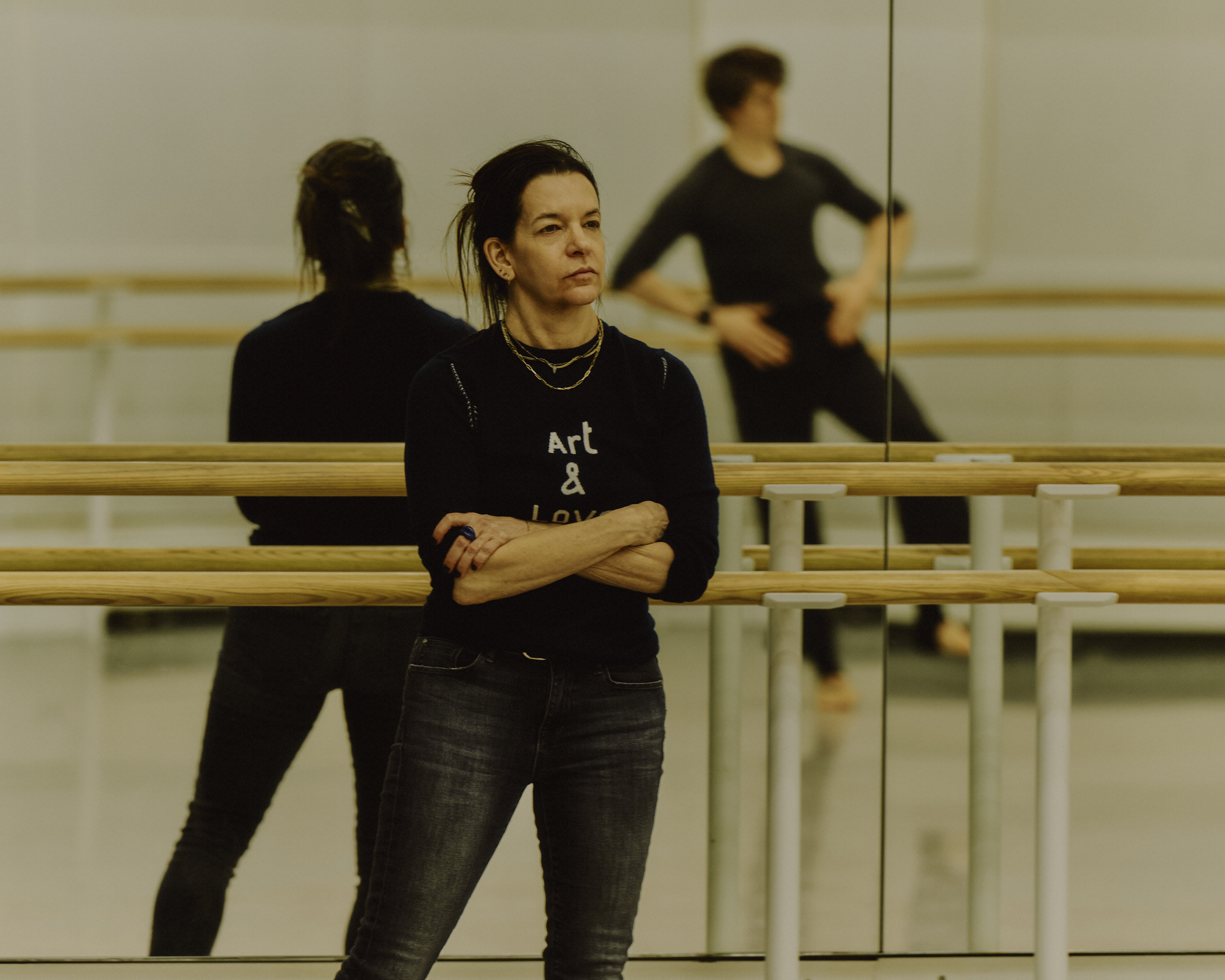 The choreographer Pam Tonowitz stands against a mirrored barre in a dance studio watching The Royal Ballet dancer William Bracewell rehearse. He is only visible in the reflection of the mirror behind where Tanowitz is standing. (1 of 4)