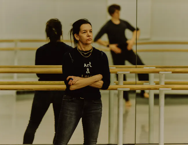 The choreographer Pam Tonowitz stands against a mirrored barre in a dance studio watching The Royal Ballet dancer William Bracewell rehearse. He is only visible in the reflection of the mirror behind where Tanowitz is standing. (1 of 4)