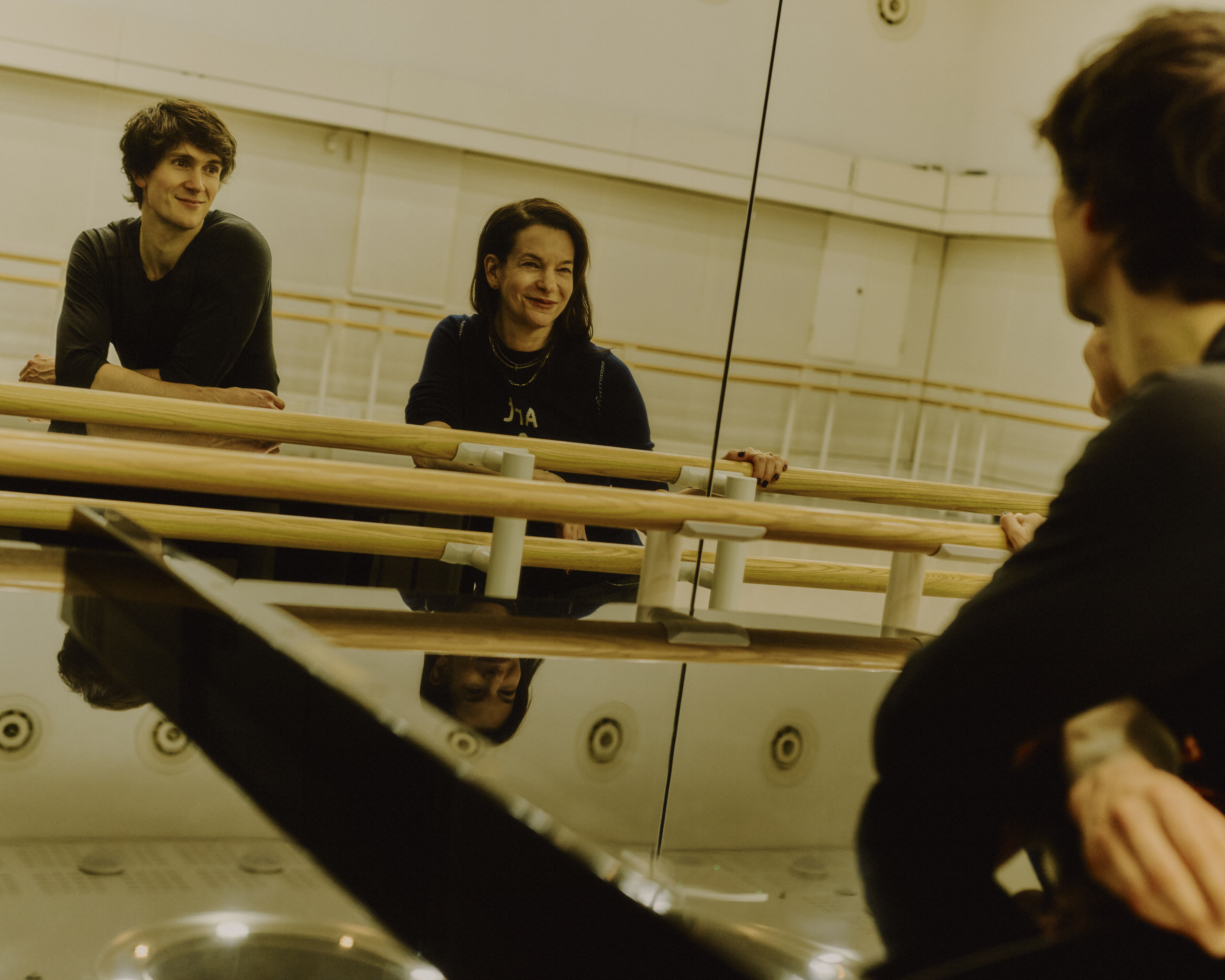 The choreographer Pam Tanowitz and The Royal Ballet dancer William Bracewell sit facing a mirrored wall against a barre in a well-lit dance studio. 