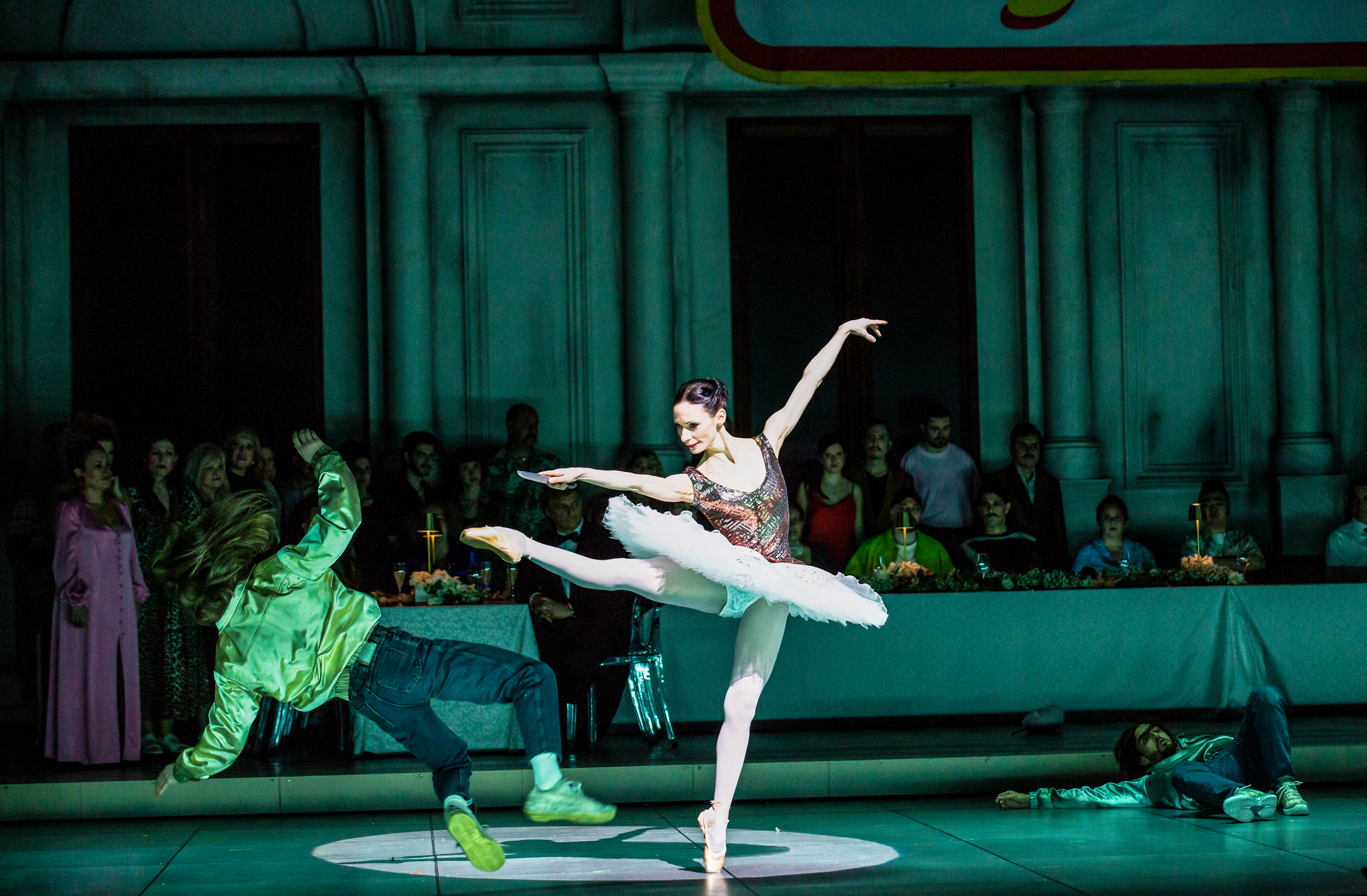 A ballet dancer wearing a tutu stands en pointe kicking out with her right leg as another performer falls to the ground. (1 of 6)