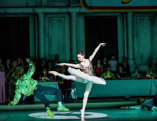 A ballet dancer wearing a tutu stands en pointe kicking out with her right leg as another performer falls to the ground. (1 of 6)