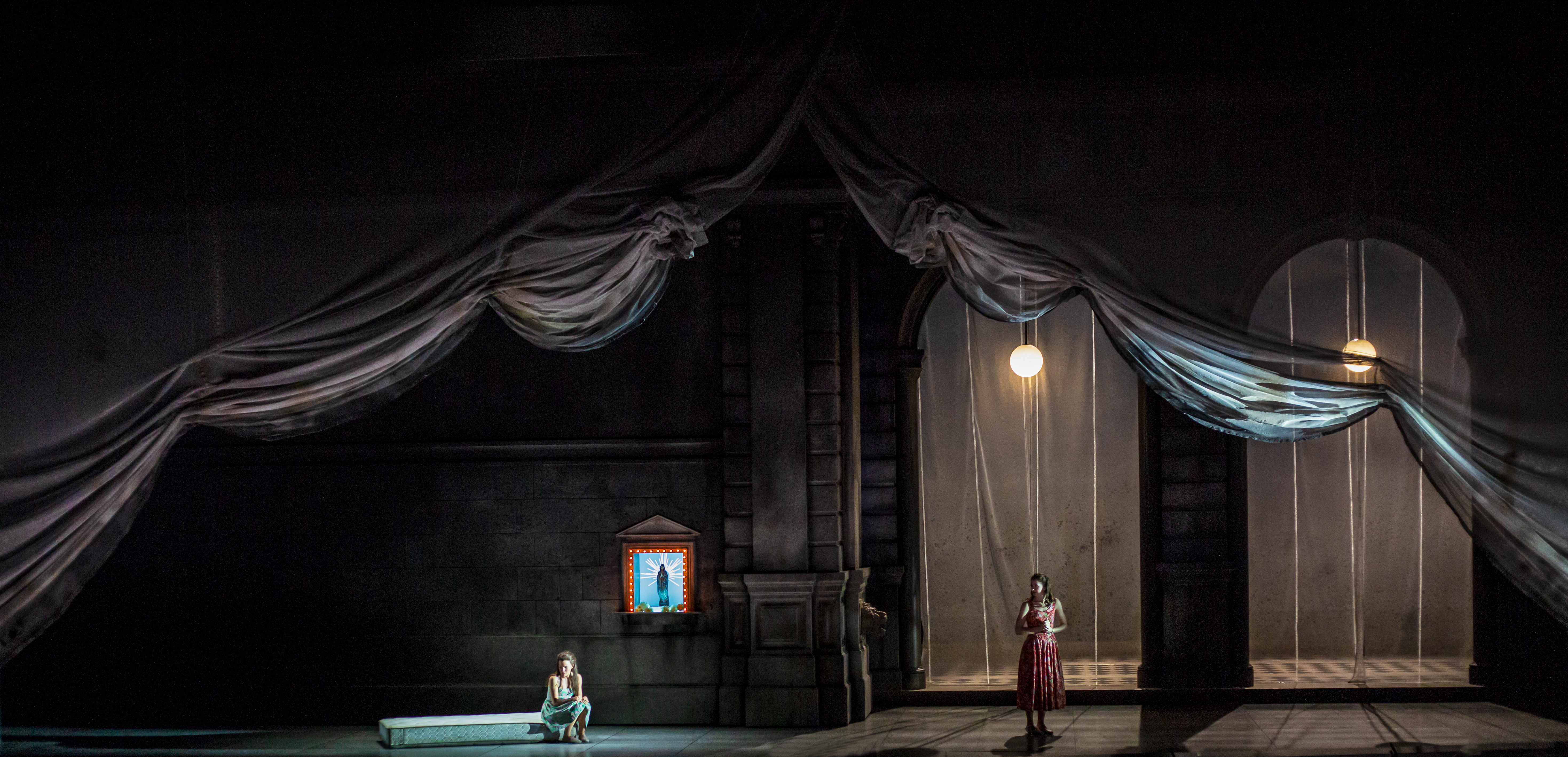 Two performers stand on a stage set with two arched doorways and a curtain draped across the whole set. One of the performers sits on a bench with a traditional church board affixed to the wall behind. (2 of 6)