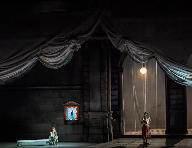 Two performers stand on a stage set with two arched doorways and a curtain draped across the whole set. One of the performers sits on a bench with a traditional church board affixed to the wall behind. (2 of 6)