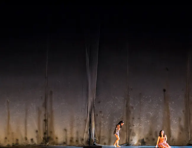 Two performers stand on dimly lit stage with a stark concrete backdrop. (3 of 6)
