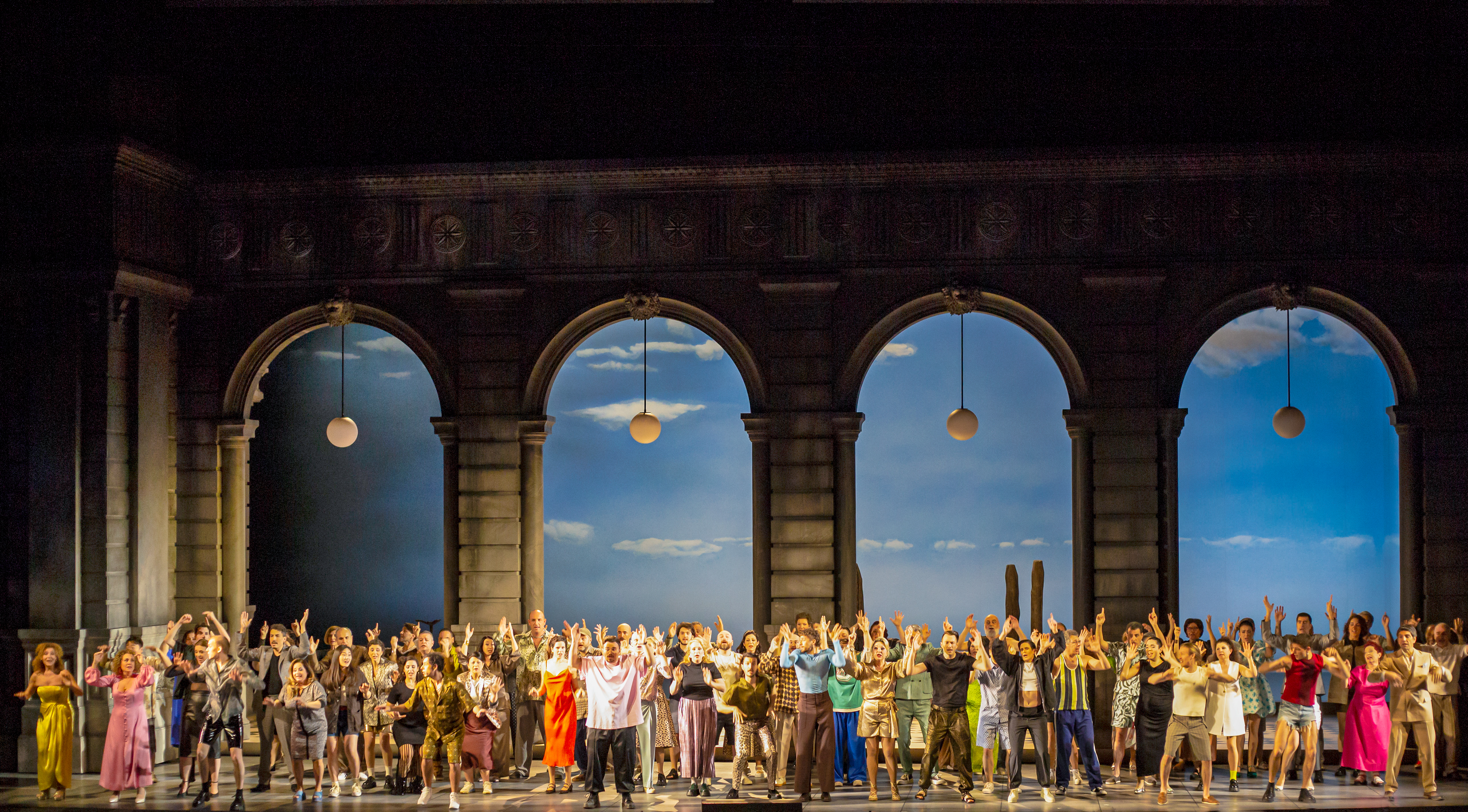 Many performers stand on stage with their arms raised high. They stand against four tall archways with street lamps hanging down and the backdrop is painted like a bright sky on a sunny day. (4 of 6)