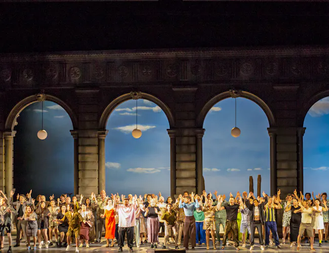 Many performers stand on stage with their arms raised high. They stand against four tall archways with street lamps hanging down and the backdrop is painted like a bright sky on a sunny day. (4 of 6)